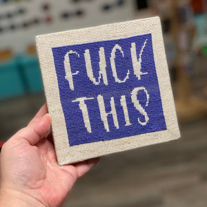 A cross stitch kit that says "Fuck This" Stitched in dark purple on an off-white canvas.