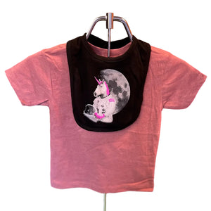 A toddler's bib shown over a dusty pink shirt and displayed on a metal hanger stand. The bib features a graphic of a white astronaut unicorn with a pink horn and wearing pink roller skates and sitting in front of a gray full moon.