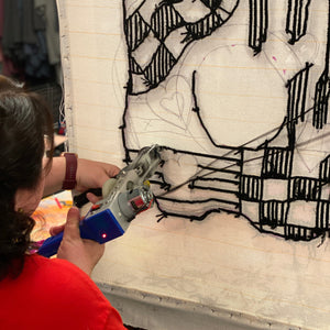 Person using a tufting gun on a piece of canvas with black line art.