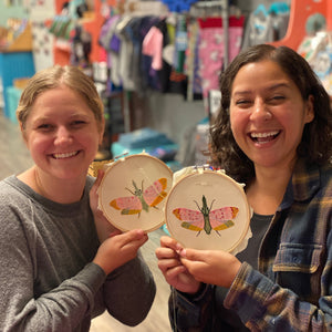 Two women holding embroidered hoop art with moth designs in a store setting.