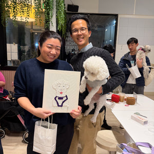 Two people holding a small dog and a punch needle portrait of that dog in a room with other people and items. The dog is looking at the portrait as if to say, "Damn! That's GOOD!"