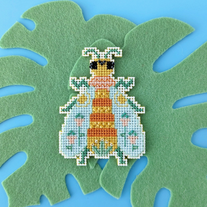 A cross stitch ornament of a bee is sitting atop a felt Monstera leaf.