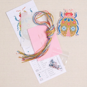 A photo of what is included in your Beetle Cross Stitch kit - floss, needles, aida fabric, a paper pattern and a bag of beads.