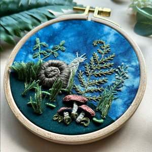 Embroidery hoop with embroidered scene of plants and a snail on blue fabric.