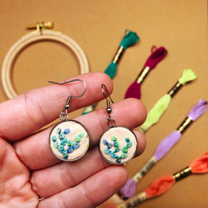 Hand holding embroidered earrings with colorful thread spools in the background.