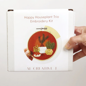 Hand holding a 'Happy Houseplant Trio Embroidery Kit' packaging on a white background.