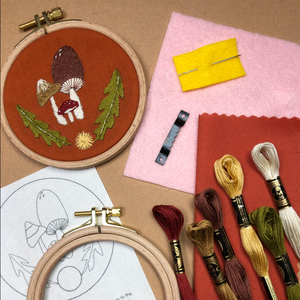 Embroidery hoop with mushroom design, fabric samples, and embroidery threads on a brown surface.