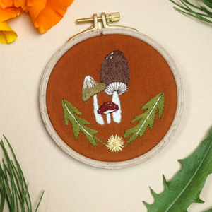 Embroidery hoop with embroidered mushrooms and leaves on a beige background with flowers and leaves.