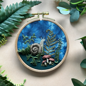 Embroidery hoop with nature-themed design surrounded by green leaves on a beige background