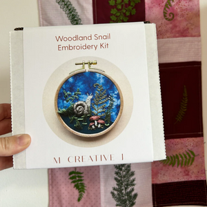 Woodland Snail Embroidery Kit packaging held by a hand with a patterned fabric background.
