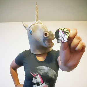 A person wearing a realistic gray unicorn mask and a black graphic t-shirt with a unicorn print, holding up a small enamel pin against a plain light gray wall.