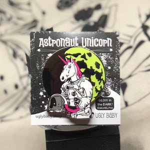 An enamel pin of an astronaut unicorn on its original backing card, photographed against a black and white patterned background.
