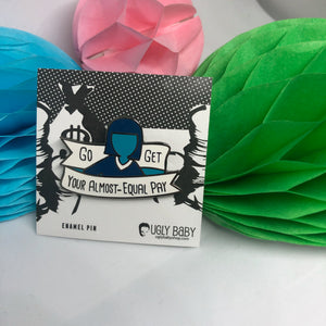 An enamel pin on its backing card, displayed in front of colorful paper honeycomb decorations. The pin features a stylized illustration of a person with a blue silhouette and black hair, holding a banner that reads 'Go GET Your ALMOST-EQUAL PAY'.