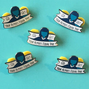 Five enamel pins featuring a stylized person with dark hair and a blue silhouette, with a banner reading 'GO GET YOUR ALMOST-EQUAL PAY', arranged on a turquoise background.