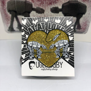 Heart shaped enamel pin called Love AT-AT First Sight with a gold glitter background. The original Shower Art piece that this design is based on is shown in the background.