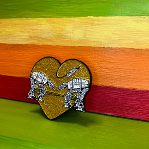Heart shaped enamel pin called Love AT-AT First Sight with a gold glitter background. Shown against a rainbow background.