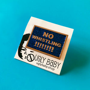 A black with rainbow glitter enamel pin that says "No Whistling !!!!!!!!" in gold text.