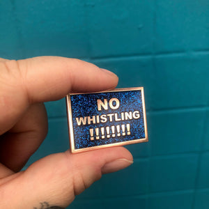 A black with rainbow glitter enamel pin that says "No Whistling !!!!!!!!" in gold text.