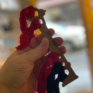 A human hand holding up an embroidery floss holder made out of cardboard. It is in the shape of the Space Needle. It has five round holes that have red and black embroidery floss in them.