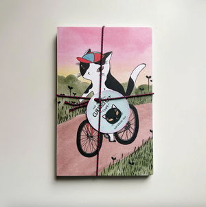 Notebook with a cat riding a bicycle illustration on the cover, tied with a string, on a white background