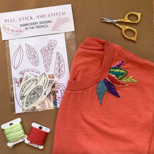 Embroidery kit with designs, thread, scissors, and embroidered shirt on a brown background.