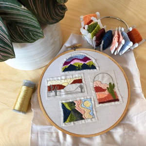 Embroidery hoop with colorful stitched landscape design on a wooden surface with thread spools.