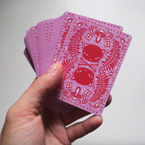 Hand holding pink playing cards with red cat designs on a light gray background.