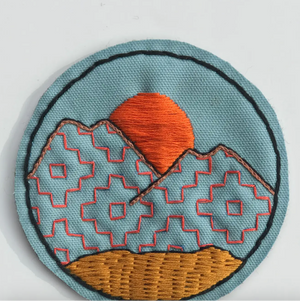 Embroidered patch with mountain and sun design on a light blue background.