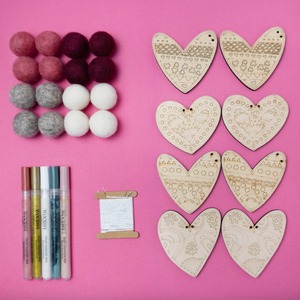 Heart-shaped wooden cutouts, felt balls, fabric markers, and thread on a pink background