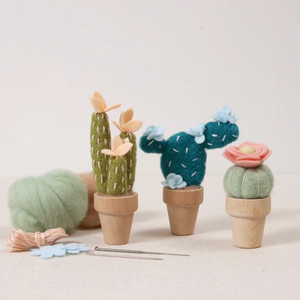 Set of needle felted cacti with wooden pots on a light background. Needle felting supplies are scattered on the table.