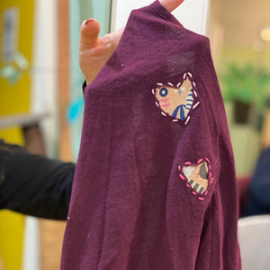 Purple sweater with heart-shaped reverse applique designs held by a hand against a blurred indoor background.