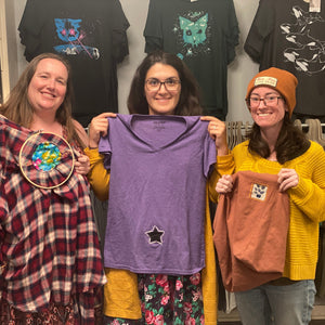 Three women holding up t-shirts in a store with clothing racks in the background.