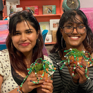 Two women holding beaded plant sculptures in a store setting.