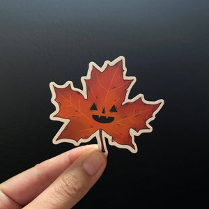 Hand holding a red maple leaf with a jack-o'-lantern face design against a black background