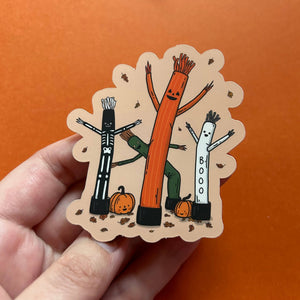 A hand holds a scalloped die-cut sticker featuring three Halloween-themed stick figures: a skeleton, a tall orange tree-like figure, and a white figure with 'BOO' written on it, with small pumpkins at the bottom. The background is a solid orange surface.