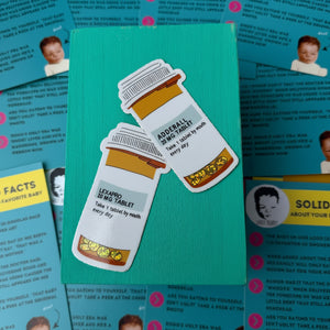 Two prescription pill bottles with labels on a teal card, surrounded by printed materials by gift shop - Ugly Baby.