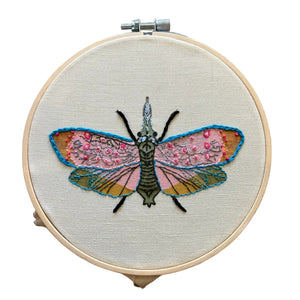 A circular embroidery hoop holding cream fabric with a detailed embroidered moth design. The moth has pink wings with small embellishments, blue outlines, and a dark green body.