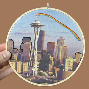 Embroidery hoop with a cityscape including the Space Needle and rainbow on a brown background.