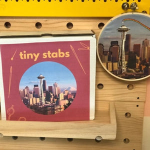 Product display with 'tiny stabs' packaging and an embroidery hoop featuring a Seattle cityscape on a pegboard.