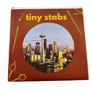 A square product box for 'Tiny Stabs' souvenir Seattle embroidery kit. The box features a terracotta red background with yellow text and line-art illustrations of sewing tools, with a circular cutout showing a photo of the Seattle skyline featuring the Space Needle.