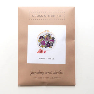The packaging for the Violet Vibes cross stitch kit is a kraft brown envelope that says "Cross stitch kit" at the top. It has a picture of the finished kit on the front.