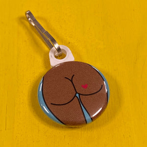 Round zipper pull of a butt with a heart tattoo on a yellow background.