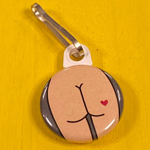 Small zipper pull with a butt design with a heart tattoo on a yellow background.