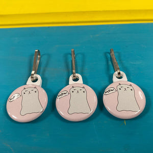 Three pink zipper pulls with cat illustrations on a blue surface.