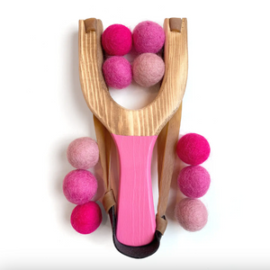 A wooden slingshot toy with a bright pink handle and several pink felt balls, photographed on a clean white background.