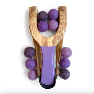 A wooden slingshot toy with a purple handle and elastic bands, with purple and lavender felt balls, photographed on a clean white background.