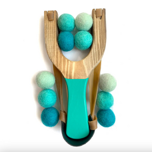 A wooden slingshot toy with a natural wood body, turquoise painted handle and rubber bands, and felted wool balls in teal, aqua, and mint green, photographed on a white background.