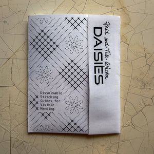 A white booklet or guidebook titled 'DAISIES' with black text and stitched geometric patterns, photographed on a textured beige surface.