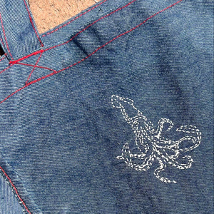 Close-up of a blue denim tote bag showing the fabric texture, red contrast stitching, and a white embroidered squid design.