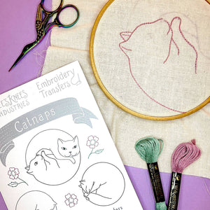Embroidery hoop with cat design, scissors, and embroidery thread on a purple surface.